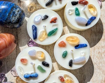 May include: Several white, circular candles are adorned with a variety of colourful gemstones. Polished stones, such as blue lapis lazuli and amethyst, are scattered around the candles. The gemstones are in shades of blue, green, yellow, red, and black.