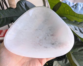 May include: A white selenite bowl with a smooth, polished surface. The bowl is shaped like a teardrop and has a slight curve to the bottom. It is a natural crystal and has a unique, organic shape.