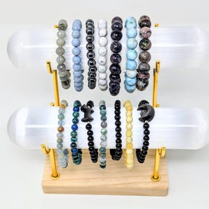 May include: A two-tiered display stand with a light wood base and gold metal supports holding two horizontal white cylindrical bars. Fourteen beaded bracelets in various colours, textures, and stone types are arranged on the bars.