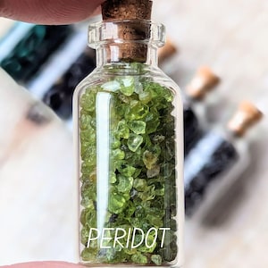 Peut inclure: Petite bouteille en verre remplie de pierres précieuses péridot vert vif. La bouteille a un bouchon en liège et le mot "PERIDOT" est imprimé sur le devant. D'autres bouteilles sont visibles en arrière-plan.