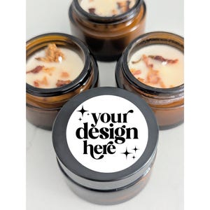 May include: Four small, brown glass jars filled with cream-coloured wax and decorative dried petals. One jar has a black lid with the text "your design here" in a bold, modern font. The jars are arranged on a white surface.