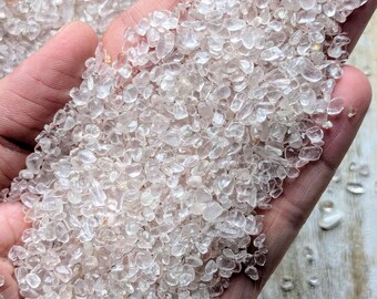 May include: A close-up of a hand holding a pile of small, clear quartz crystals. The crystals are tumbled and have a smooth, polished finish.