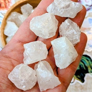 May include: A selection of rough, clear quartz crystals. The crystals are held in a hand, with a wooden bowl of similar crystals visible in the background. Colourful stickers are also in the background.