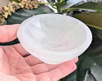 May include: A white selenite bowl with a smooth, polished surface. The bowl is slightly curved and has a rounded bottom.