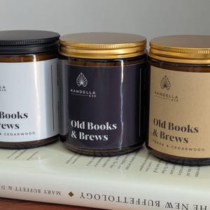 May include: Three glass jar candles with lids, each labeled "Old Books & Brews." The jars have different colored labels: white, dark blue, and brown. The candles sit on a book.