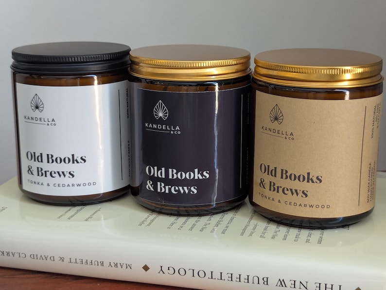 May include: Three glass jar candles with lids, each labeled "Old Books & Brews." The jars have different colored labels: white, dark blue, and brown. The candles sit on a book.