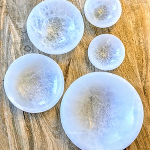 May include: A collection of four circular bowls crafted from translucent white selenite. Each bowl features a smooth, polished surface with visible natural crystalline textures and internal fractures, varying in size from small to large.