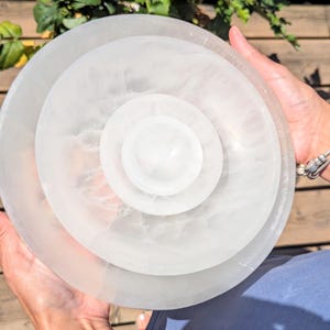 May include: A circular, translucent white selenite bowl featuring a smooth, polished surface with natural cloudy veining. The bowl has a tiered, concentric design with a smaller, raised circular centre section nested within a wider, shallow basin.