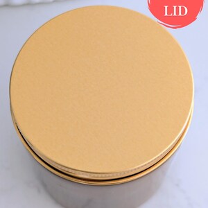 May include: A round container with a gold lid. The lid is labeled "GOLD LID" in a red circle. The container's body is a dark, muted color, and the lid appears to be made of metal. The container sits on a white surface.