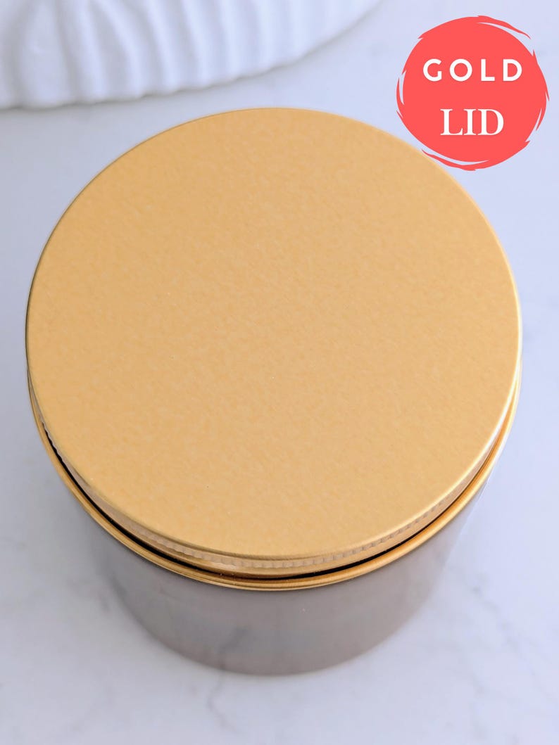 May include: A round container with a gold lid. The lid is labeled "GOLD LID" in a red circle. The container's body is a dark, muted color, and the lid appears to be made of metal. The container sits on a white surface.