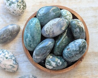 May include: A bowl of green, gray, and white moss agate tumbled stones. The stones are smooth and polished, and they have a variety of patterns and textures.
