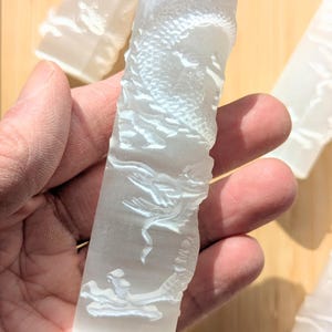 Puede incluir: Una varita de cristal transparente y rectangular con un diseño de dragón tallado. El dragón está intrincadamente detallado, con escamas y elementos fluidos. Se ven otras varitas similares en el fondo.