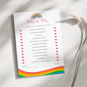 Puede incluir: Una hoja de juego blanca con el texto "Mrs & Mrs" y un arcoíris. La hoja incluye preguntas como "¿Dijo te amo primero?" y "¿Es más divertido?" con espacios para respuestas. Gráficos de labios rojos están a los lados.
