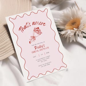 Könnte beinhalten: Eine rosa und weiße Einladung zur Brautparty mit rotem Rand und einer roten Blumenillustration. Der Text lautet "That's amore" und "Ruby's Bridal Shower". Die Einladung enthält Datum, Uhrzeit und Ort der Veranstaltung sowie die RSVP-Informationen.