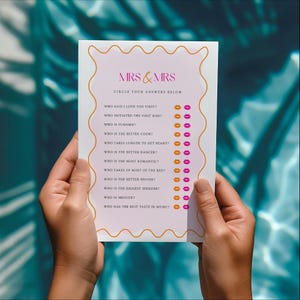 May include: A white card with the text "MRS & MRS" in pink and a decorative orange border. The card features a list of questions with pink and orange lip icons. The card is held by two hands.