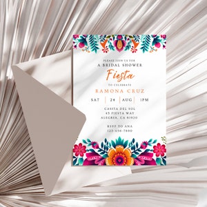 Op de afbeelding: Een witte uitnodiging met kleurrijke bloemmotieven. De tekst luidt "Please join us for a bridal shower Fiesta to celebrate Ramona Cruz Za 24 aug 13:00 Casita del Sol 45 Fiesta Way Alegria, CA 81920 RSVP aan Ana 123-456-7890"