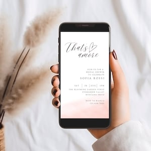 May include: A pink and white digital invitation for a bridal shower. The invitation reads "That's amore" with a heart symbol and includes the details for the event: "Join us for a bridal shower to celebrate Sofia Rossi, Sat 24 Aug 1PM, The Blooming Garden Venue, Evergreen Valley, Montana 09070, RSVP to Bianca 123-456-7890."