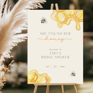 Op de afbeelding: Een beige bruiloftsshowerbord met een aquarelillustratie van honingraten en bijen. De tekst luidt "She found her honey" en "Welcome to Zara's Bridal Shower" met de datum "Sat 15 Mar".