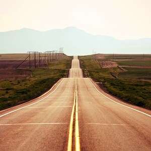 May include: A long, straight, paved road with yellow lines down the centre, leading towards a distant mountain range. The road is surrounded by green grass and fields.