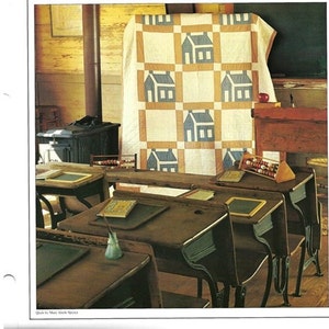 May include: A vintage classroom setting with wooden desks and a quilt hanging on the wall. The quilt is made of white and tan fabric with blue houses printed on it. The desks are arranged in rows and have small black slates on them.