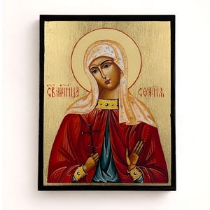 Può includere: Icona di una donna in abito rosso che tiene una croce, su sfondo dorato. L'icona ha una cornice nera e presenta testo rosso. La donna ha un'aureola e indossa un copricapo bianco.