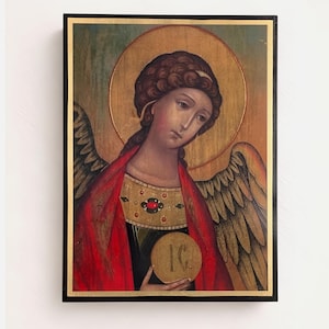 Puede incluir: Icono de un ángel con aureola dorada, vestido con una túnica roja y sosteniendo un objeto circular. El ángel tiene cabello castaño y alas emplumadas. La obra de arte está enmarcada en un marco negro con ribete dorado.