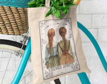 May include: A white canvas tote bag with a design of two women with long braids, wearing dresses. The text "Kindred Spirits" is printed on the bag. The bag is hanging on a bicycle handlebar with a basket filled with flowers and groceries.