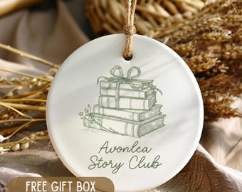 May include: A white ceramic ornament with a stack of books illustrated in green, tied with a bow. The text "Avonlea Story Club" is below the books. A brown tag at the bottom says "FREE GIFT BOX INCLUDED". A jute string is attached.