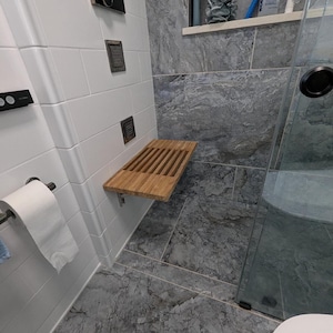 Op de afbeelding: Een rechthoekige douchebank van bamboe met latten, gemonteerd aan een wit betegelde muur. De bank heeft een natuurlijke houtnerfafwerking en wordt ondersteund door metalen beugels. Het bevindt zich in een doucheruimte met grijze marmeren tegels.