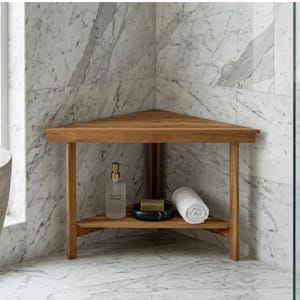 May include: A wooden corner shower bench with a shelf. The bench is made of light brown wood and has a triangular shape. A clear bottle of Argan Oil shampoo, a black soap dish, a bar of soap, and a rolled white towel are on the shelf.