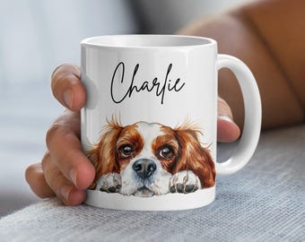 May include: White ceramic mug with a brown and white Cavalier King Charles Spaniel dog illustration. The dog is looking at the viewer with its head resting on the rim of the mug. The text "Charlie" is written in black cursive on the mug.