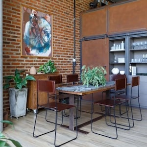 May include: A rustic dining room with a wooden table and six brown leather chairs with black metal frames. The table is set with a vase of green plants. The room has exposed brick walls and a large painting on the wall.