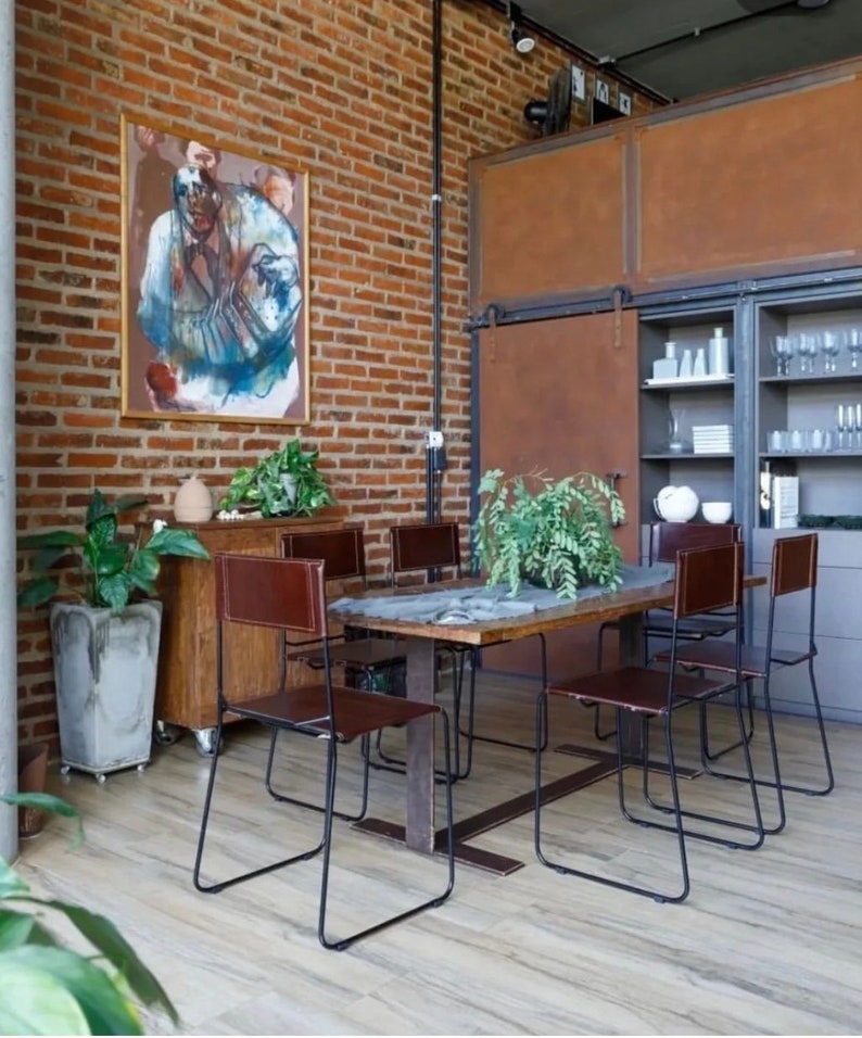 May include: A rustic dining room with a wooden table and six brown leather chairs with black metal frames. The table is set with a vase of green plants. The room has exposed brick walls and a large painting on the wall.