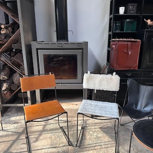 May include: Two chairs with black metal frames. One chair has a tan leather seat and back. The other chair has a white fluffy seat and back. A fireplace and shelves are in the background.