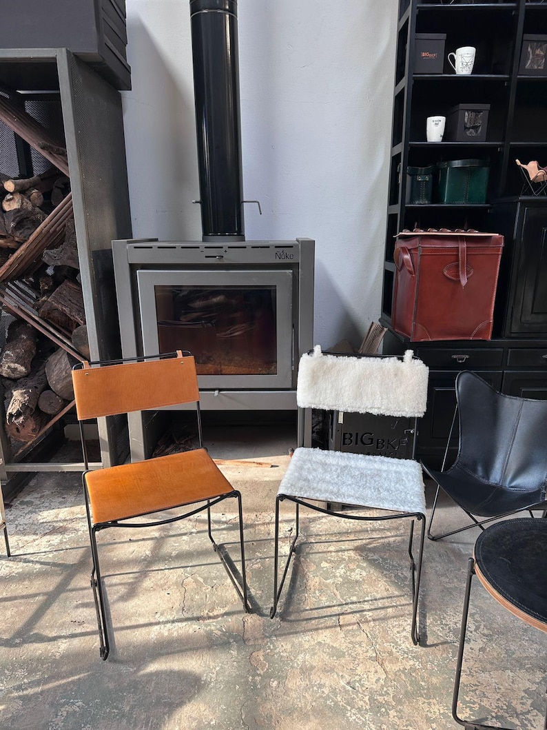 May include: Two chairs with black metal frames. One chair has a tan leather seat and back. The other chair has a white fluffy seat and back. A fireplace and shelves are in the background.
