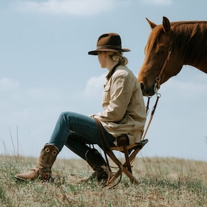 May include: A woman wearing a brown hat and a beige jacket sits on a wooden folding chair in a field. A brown horse stands next to her, its head turned towards her.