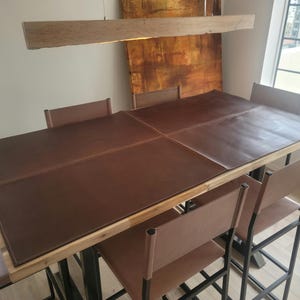 Puede incluir: Un camino de mesa de cuero marrón con bordes cosidos se encuentra sobre una mesa de madera con patas de metal negro y sillas de cuero marrón.