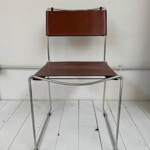 May include: A brown leather chair with a rectangular back and seat. The chair has a chrome frame and a minimalist design. The back of the chair is attached to the frame with leather straps.