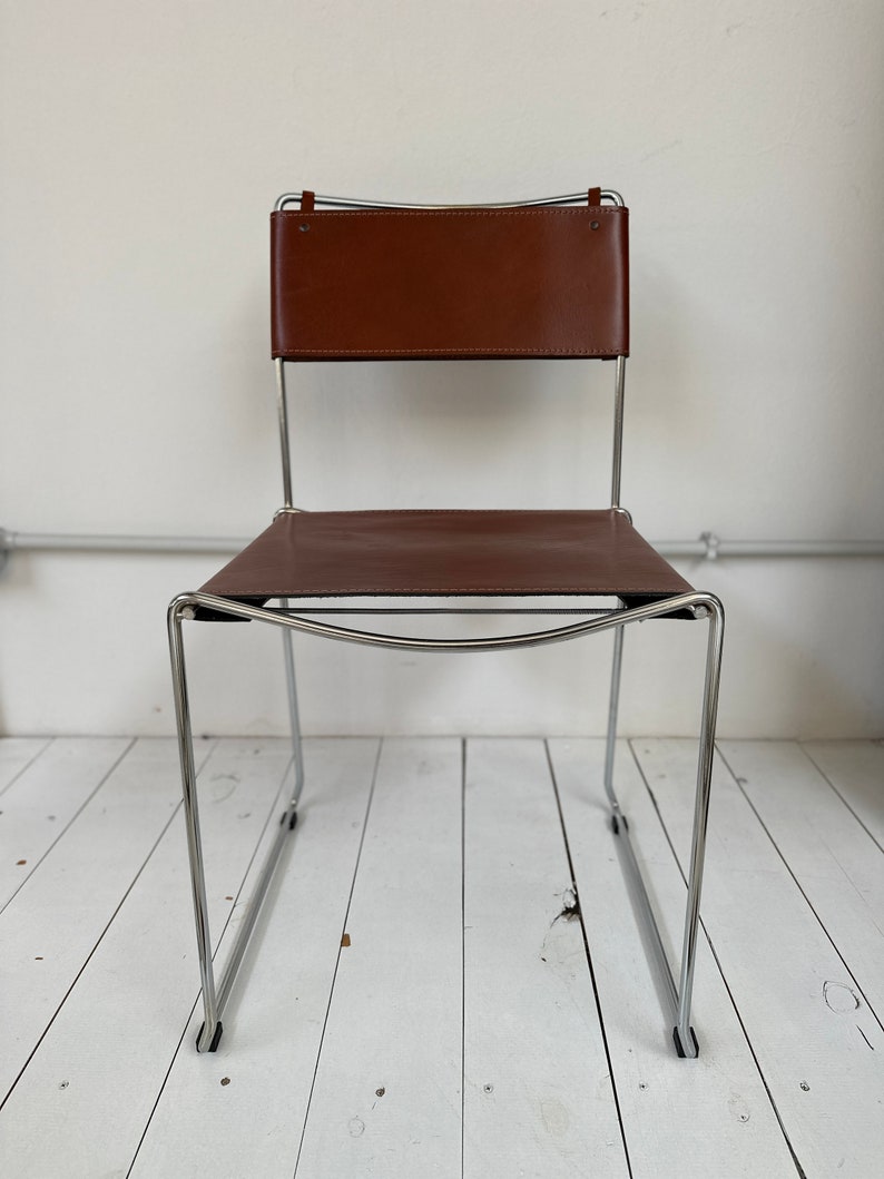 May include: A brown leather chair with a rectangular back and seat. The chair has a chrome frame and a minimalist design. The back of the chair is attached to the frame with leather straps.