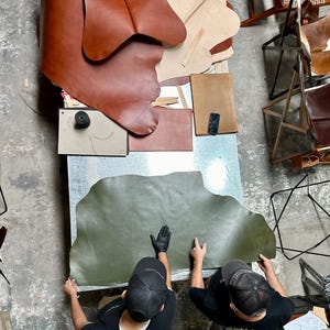 May include: Overhead shot of a workshop with leather hides and tools. Two people wearing black shirts and caps are working with a large, olive green leather hide. Various brown leather pieces and other materials are scattered on a metal table.
