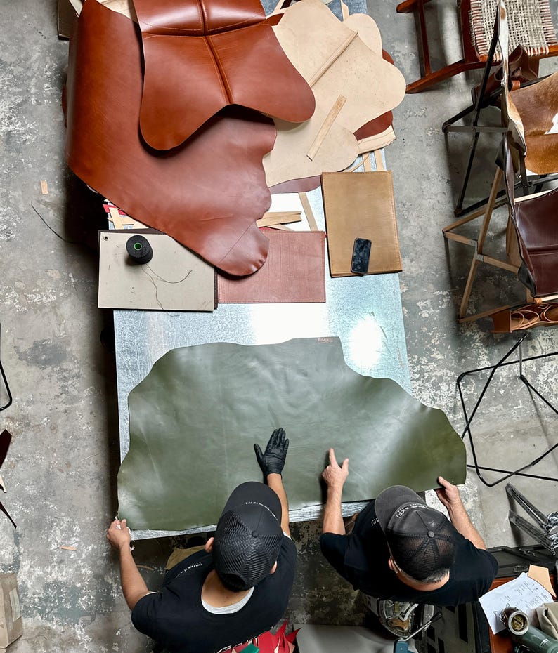 May include: Overhead shot of a workshop with leather hides and tools. Two people wearing black shirts and caps are working with a large, olive green leather hide. Various brown leather pieces and other materials are scattered on a metal table.