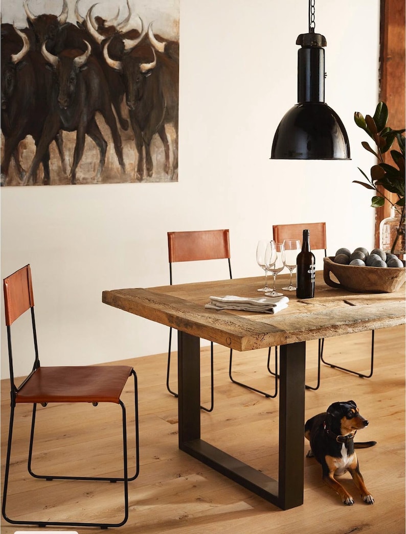 May include: A rustic wooden dining table with a black metal frame and four brown leather chairs. A black pendant light hangs above the table. A small dog is lying on the floor under the table.