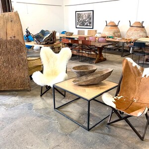 May include: Two chairs, one with a white sheepskin cover and the other with a brown and white cowhide cover, are arranged around a square wooden coffee table with a black metal frame. The chairs are in a large, open room with a concrete floor. There are wooden bowls on the table and other furniture in the background.