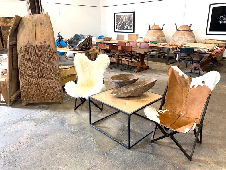 May include: Two chairs, one with a white sheepskin cover and the other with a brown and white cowhide cover, are arranged around a square wooden coffee table with a black metal frame. The chairs are in a large, open room with a concrete floor. There are wooden bowls on the table and other furniture in the background.