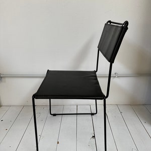 May include: Black leather chair with a minimalist design. The chair features a rectangular seat and backrest, supported by a black metal frame. The chair is positioned against a white wall and white wooden floor.