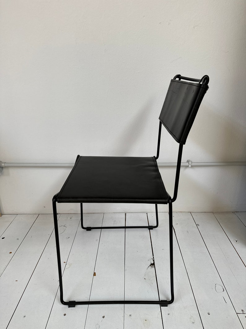 May include: Black leather chair with a minimalist design. The chair features a rectangular seat and backrest, supported by a black metal frame. The chair is positioned against a white wall and white wooden floor.