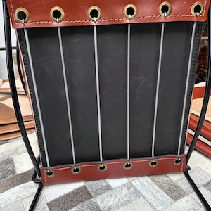 May include: A handcrafted magazine rack with a black metal frame. The rack features a black interior and is trimmed with brown leather at the top and bottom, secured with brass grommets. Vertical metal rods provide support. The rack is designed for storing magazines.