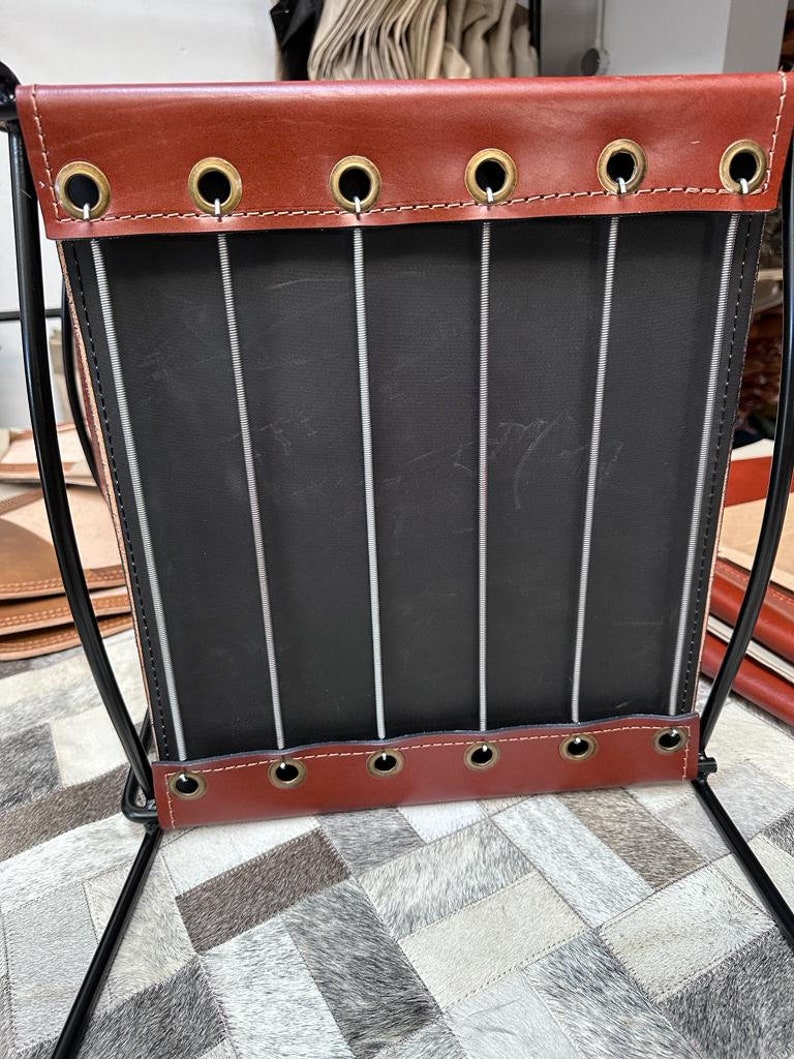 May include: A handcrafted magazine rack with a black metal frame. The rack features a black interior and is trimmed with brown leather at the top and bottom, secured with brass grommets. Vertical metal rods provide support. The rack is designed for storing magazines.