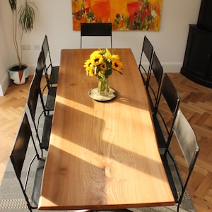 May include: A long wooden dining table with a natural wood finish and a black metal base. The table is surrounded by six black metal chairs with leather seats. A vase of yellow sunflowers sits in the center of the table.
