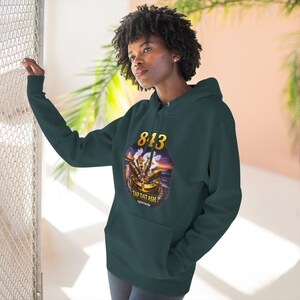 May include: A dark teal hoodie with a front pocket and a drawstring hood. The front features a gold-colored oval design with the number "813" and the text "TAP DAT ASH" in white. The design includes an illustration of a smoking object.